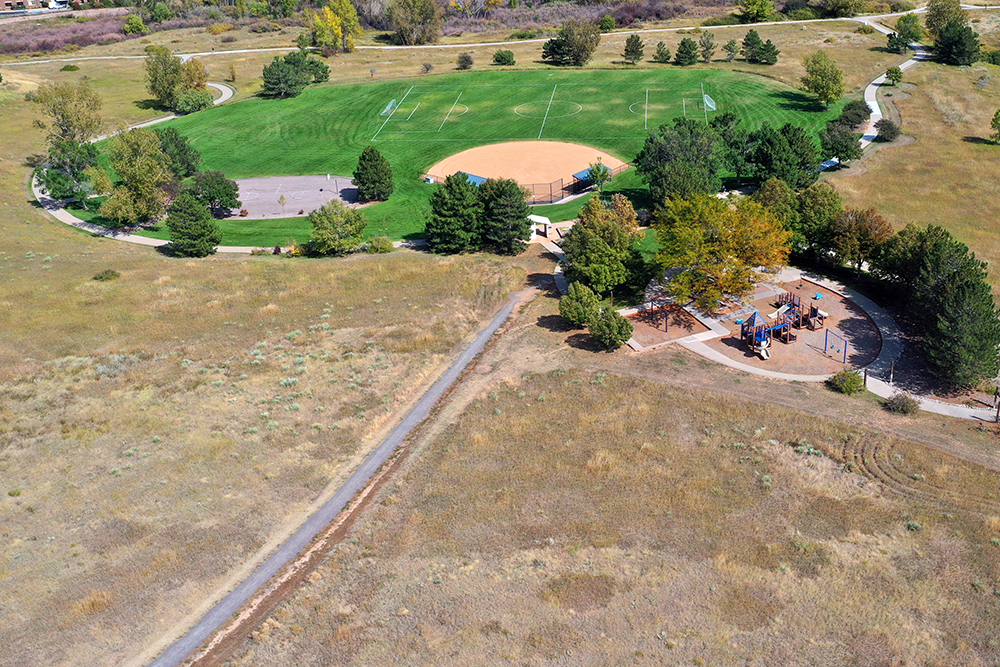 An aerial view of Toepfer Park with an open natural grass field in the foreground, a playground and tall shade trees, and a basketball court near a green grassy ball field in the background.