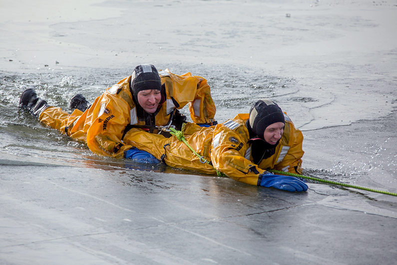 Two firefighters wearing yellow full body ice rescue suits crawl across the surface of an icy pond during a training exercise.