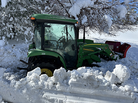 A green tractor with a plow removes heavy snow on a sidewalk