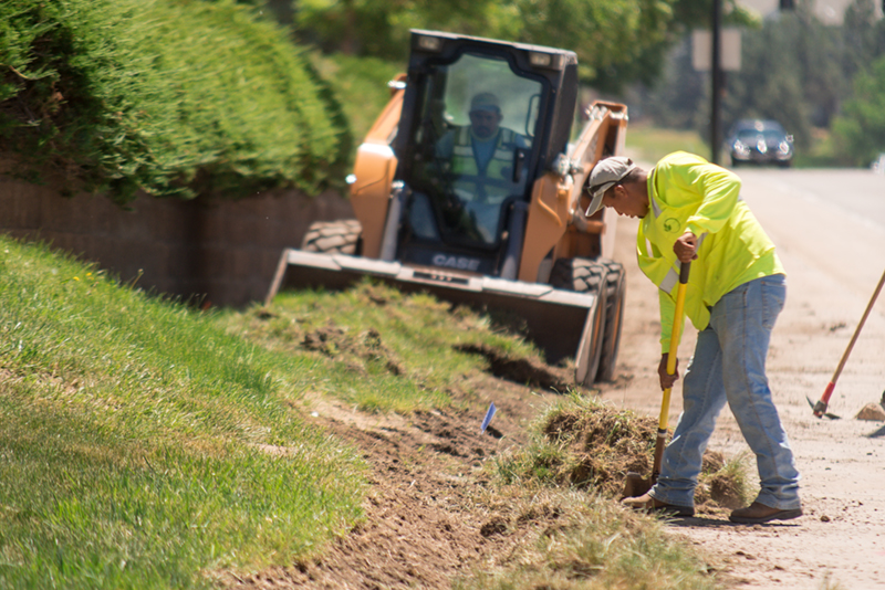 Two landscaping contractors use a bulldozer and shovel to remove landscaping along a major roadway. 