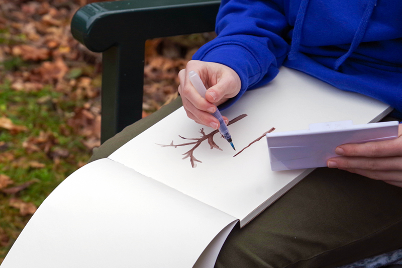 Close up of someone's hands while painting a watercolor tree in a sketchbook.